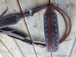 A close-up of ornate leather horse tack with detailed tooling and turquoise stone accents. The tack, consisting of straps and a buckle, is laid out on a wooden surface.