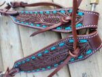 A close-up of intricately designed leather straps lying on a wooden surface. The brown leather features carved leaf patterns and turquoise accents, highlighted by white stitching along the edges. Thin leather ties and metal hardware are also visible.