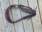 A brown leather horse breast collar with intricate blue and black stitching details, silver buckles, and leather straps is laid out on a wooden surface. The collar features decorative floral patterns and is designed for attachment to a horse's tack.