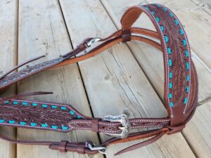 An intricately designed leather horse breast collar rests on a wooden surface. The brown leather is detailed with turquoise accents and white stitching, featuring carved feather patterns and a decorative silver buckle.