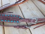 A close-up of a leather horse bridle adorned with intricate floral patterns and turquoise accents. The image shows a detailed view of the tooling and stitching, with a silver buckle in the center. The bridle lies on a wooden surface.