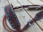 A detailed image of a handmade leather horse breast collar with intricate floral tooling and turquoise accents. The piece is displayed on a wooden surface, showcasing its fine craftsmanship and silver buckle.