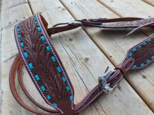 A detailed image of a handmade leather horse breast collar with intricate floral tooling and turquoise accents. The piece is displayed on a wooden surface, showcasing its fine craftsmanship and silver buckle.