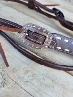 A close-up of a brown leather belt with an ornate, silver-colored buckle accented with small, round embellishments. The belt is lying on a wooden surface, showcasing stitching details and leather straps.