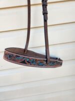 A close-up photo of a leather belt with an engraved design featuring blue leaves and swirls. The belt is brown with white stitching along the edges and is displayed hanging against a beige horizontal paneled background.