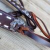 Close-up of a piece of dark brown leather with decorative stitching and a silver floral concho. The leather is looped through the concho and tied with a thin strip, laying on a wooden surface in the background.