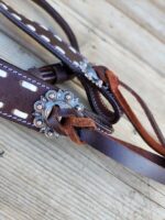 Close-up of a piece of dark brown leather with decorative stitching and a silver floral concho. The leather is looped through the concho and tied with a thin strip, laying on a wooden surface in the background.