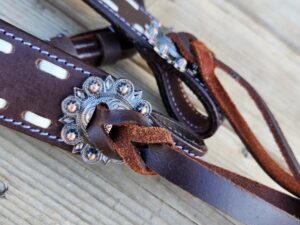 Close-up of a piece of dark brown leather with decorative stitching and a silver floral concho. The leather is looped through the concho and tied with a thin strip, laying on a wooden surface in the background.