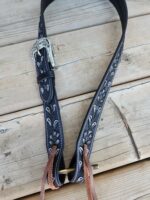 A black leather guitar strap with white embroidered patterns and a silver buckle. The strap is placed on a wooden surface and has leather lace-like ties at the end.