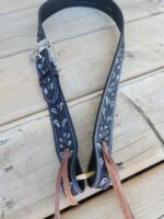 A decorative black leather guitar strap with white stitching and intricate silver floral patterns is laid on a wooden surface. The strap features a silver buckle and two brown leather strings attached at one end.