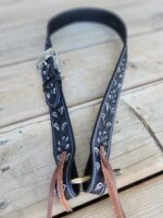 A decorative black leather guitar strap with intricate white stitching and a silver buckle, lying on a wooden surface. The ends of the strap have brown leather laces for attaching to a guitar.