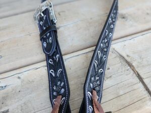 A decorative black leather guitar strap with intricate white stitching and a silver buckle, lying on a wooden surface. The ends of the strap have brown leather laces for attaching to a guitar.