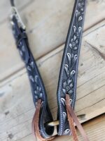 A close-up image of a dark leather guitar strap resting on a wooden surface. The strap features intricate floral designs with white accents and has leather ties at the end. The background shows the texture of the wooden surface.