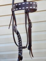 Close-up of a brown leather horse bridle with white stitching, hanging against a beige corrugated background. The bridle includes detailed silver buckles and straps.