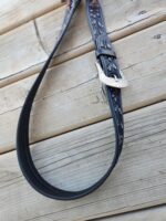 Close-up image of a black leather belt with intricate embossed patterns hanging over a wooden surface. The belt has a shiny, ornate metal buckle that contrasts with the rustic backdrop.