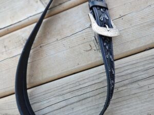 Close-up image of a black leather belt with intricate embossed patterns hanging over a wooden surface. The belt has a shiny, ornate metal buckle that contrasts with the rustic backdrop.