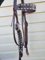 A brown leather horse bridle with white stitching, featuring decorative metal buckles and accents, is hanging against a beige, horizontally paneled background. The bridle includes reins and a decorated browband.