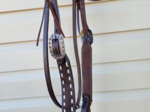 A brown leather horse bridle with white stitching, featuring decorative metal buckles and accents, is hanging against a beige, horizontally paneled background. The bridle includes reins and a decorated browband.