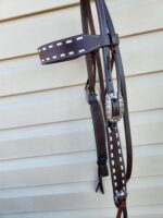 Close-up of a dark brown leather bridle with white stitching and silver embellishments, hanging against a light-colored, horizontally slatted surface. The bridle includes various straps and a decorative buckle.