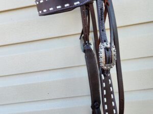 Close-up of a dark brown leather bridle with white stitching and silver embellishments, hanging against a light-colored, horizontally slatted surface. The bridle includes various straps and a decorative buckle.