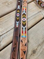 Two leather belts lie side by side on a wooden surface. Both feature intricate beadwork with geometric patterns in vibrant colors including red, yellow, white, and black. The belts also have detailed carved designs on the leather sections.