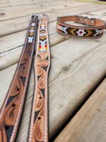 A close-up of two leather belts and a leather collar on a wooden surface. The belts feature intricate tooled designs and colorful beadwork patterns, while the collar also has detailed beadwork with geometric shapes.