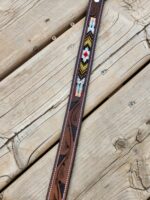 A close-up of a decorative leather belt with intricate patterns. The belt has colorful beadwork, including geometric and diamond designs in red, yellow, black, and white. The background is rustic wooden planks with visible grain and nails.