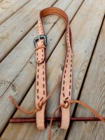 A tan leather horse bridle with red stitching lies on a wooden surface. The bridle has a silver buckle and leather strings at the ends.