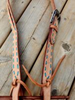 A close-up image of a leather horse bridle laid on a wooden surface. The bridle features light tan leather with turquoise stitching and a detailed metal buckle. The wood surface showcases natural textures and knots.