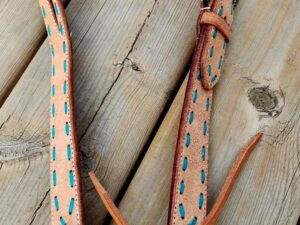 A close-up image of a leather horse bridle laid on a wooden surface. The bridle features light tan leather with turquoise stitching and a detailed metal buckle. The wood surface showcases natural textures and knots.