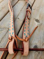 A close-up of leather reins with turquoise stitching and tied knots laying on a weathered wooden surface. The reins are adorned with a metal buckle near the top. The wood has visible grain patterns and knots, adding an element of rustic texture to the image.