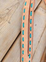 Close-up of a tan leather strap with turquoise rectangular embellishments and white stitching along the edges. The strap lies on a wooden surface with a visible knot in the wood grain.