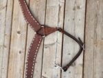 A brown leather horse harness with white stitching is laid out on a wooden surface. The harness features metal buckles and a metal clip. The wood has a rustic, weathered appearance with visible knots and grain patterns.