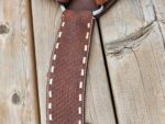 Close-up of a brown leather product with white stitching running along the sides, placed on a rustic wooden surface. The leather appears textured, with a patterned design, and the stitching is evenly spaced and parallel. The wood shows visible grain and knots.