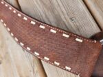 Close-up of a brown textured leather belt with white stitching along the edges, placed on a wooden surface. The belt features a woven pattern and metal buckle partially visible to the right.