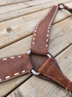 Close-up of a handcrafted leather harness with beige stitching, laid out on a wooden surface. The harness features a metal ring at the center where straps converge. The leather appears textured and slightly worn.