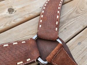 Close-up of a handcrafted leather harness with beige stitching, laid out on a wooden surface. The harness features a metal ring at the center where straps converge. The leather appears textured and slightly worn.