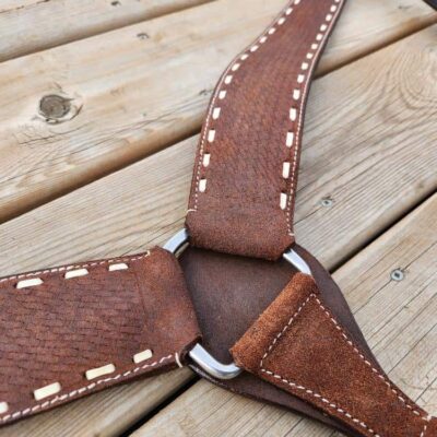 Close-up of a handcrafted leather harness with beige stitching, laid out on a wooden surface. The harness features a metal ring at the center where straps converge. The leather appears textured and slightly worn.