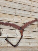 A brown leather horse breast collar with white stitching and metal hardware is laid out on a wooden surface. The collar includes two straps with metal clips and an adjustable strap at the bottom.