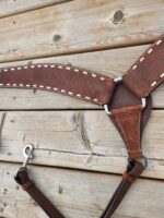 A close-up of a leather horse harness with white stitching, a metal ring, and a metal clasp. The harness is laid flat against a wooden, weathered surface, highlighting the detailed craftsmanship and texture of both the leather and the wood.