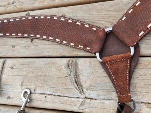 A close-up of a leather horse harness with white stitching, a metal ring, and a metal clasp. The harness is laid flat against a wooden, weathered surface, highlighting the detailed craftsmanship and texture of both the leather and the wood.