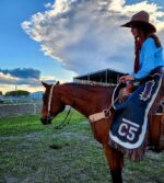 A person wearing a cowboy hat and blue shirt sits on a brown horse fitted with a Stockmans- Roughout Breast Collar- Turquoise Buckstitch at an outdoor venue. Both the rider and horse are facing left. The rider has chaps with "C5" on them. In the background, a large cloud dominates the sky above a partially visible structure.