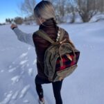 A person with long braided hair, wearing a black vest, grey sleeves, and carrying The Rocky Mountain Zip Up Backpack- Explorer Series, walks through a snowy landscape while pointing ahead. Trees and footprints are visible in the background. The intricate designs on the backpack resemble an old leather saddle blanket.