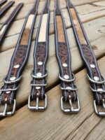 A row of intricately designed leather belts with metal buckles and D-rings, laid out on a wooden surface. The belts feature decorative stitching and tooled patterns, along with patches of lighter material.