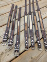 A selection of eight intricately designed leather belts with metal buckles laid out on wooden planks. The belts feature carved patterns and a combination of dark brown and beige colors. Some belts have lighter patches creating a unique, rustic appearance.