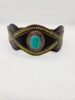 A leather bracelet with intricate green and black patterns and a central turquoise stone. The design features woven elements and decorative embellishments on the sides. The background is plain white.