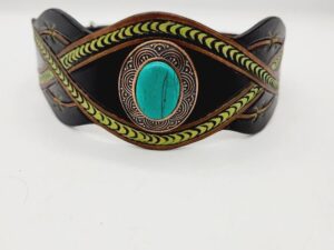 A leather bracelet with intricate green and black patterns and a central turquoise stone. The design features woven elements and decorative embellishments on the sides. The background is plain white.