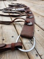 A close-up of a rustic leather breast collar with decorative metal conchos and leather ties,The breast collar features white stitching along the edges and a large metal ring at one end, giving it a handcrafted, vintage appearance.