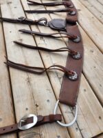 A leather horse harness with intricate silver adornments is spread out on a wooden surface. The harness features multiple straps, buckles, and decorative metal conchos, showcasing craftsmanship and attention to detail.