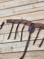 A leather horse bridle is laid out on a wooden surface. It has intricate stitching, metal rings, and straps with buckles. The bridle is designed for equestrian use, showcasing craftsmanship and durability.